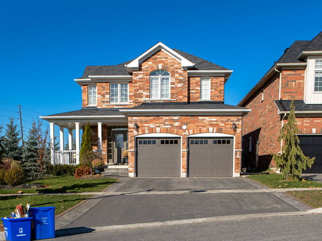 A red brick two storey detached home in a Halton subdivision, with a covered porch, a double garage and the recycling bins out at the curb